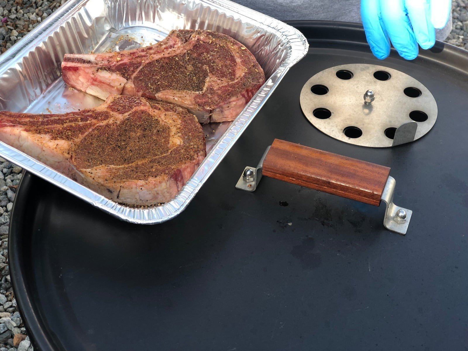 Two raw, seasoned ribeye steaks in a disposable aluminum tray next to a person wearing blue gloves, with a barbecue grill lid and a vented disc nearby.