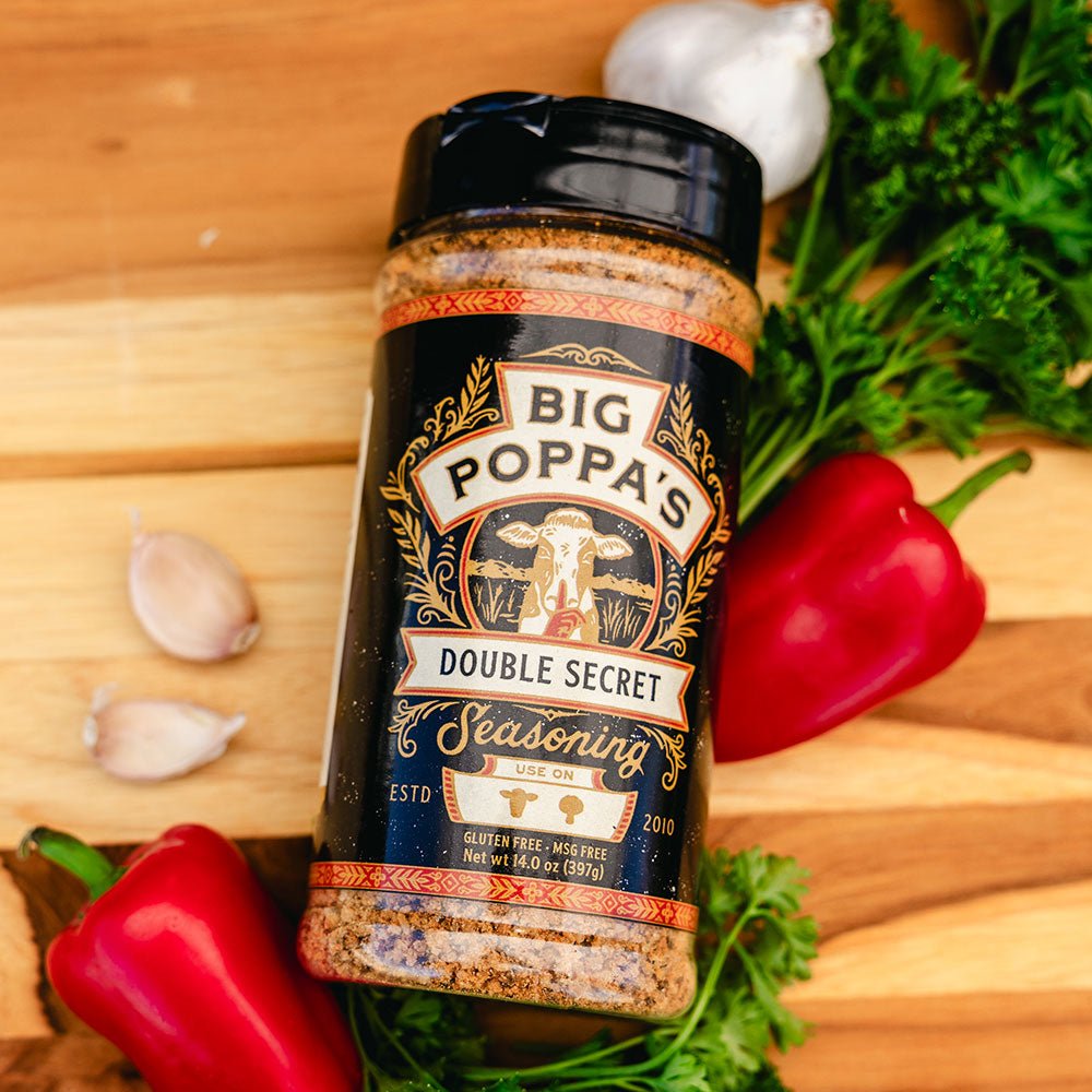 A bottle of Big Poppa's Double Secret Seasoning lying on a wooden surface, surrounded by fresh garlic cloves, red bell peppers, and parsley.