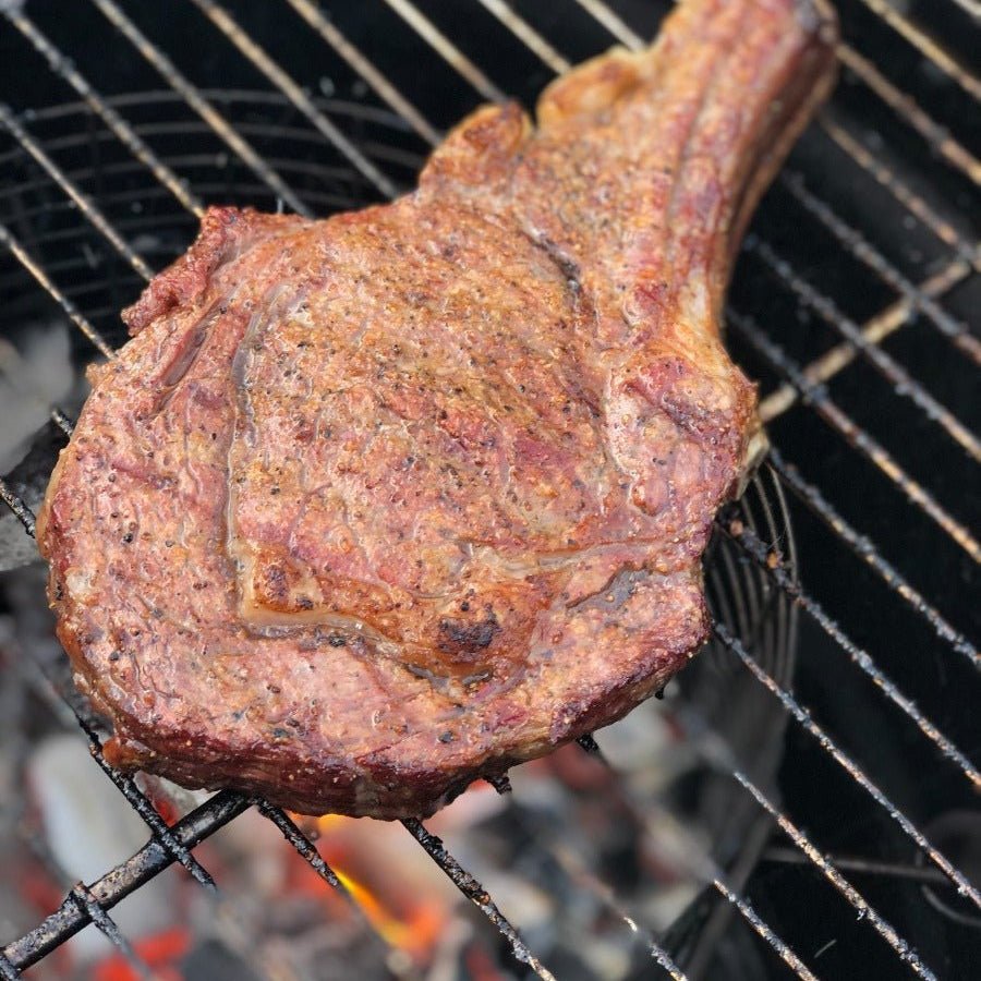 A close-up view of a large, grilled steak with a well-browned, seasoned crust, cooking over glowing charcoal on a barbecue grill.