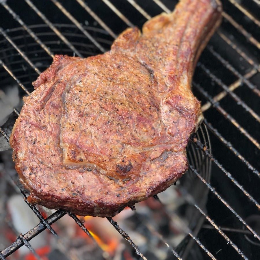 A close-up view of a large, grilled steak with a well-browned, seasoned crust, cooking over glowing charcoal on a barbecue grill.