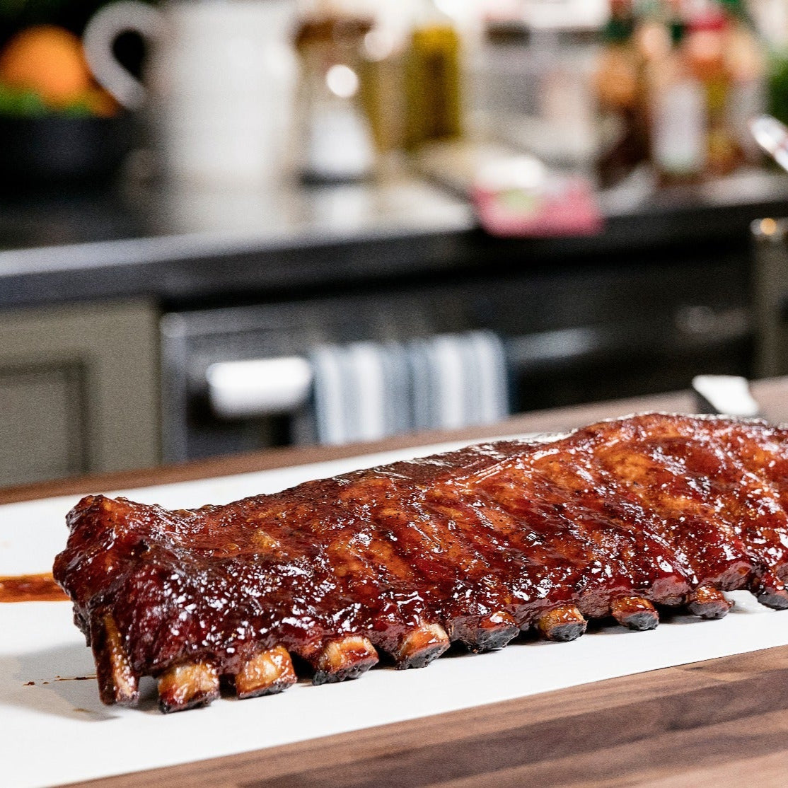 Glazed barbecue ribs placed on a white serving board in a modern kitchen setting. The ribs are richly coated in a shiny barbecue sauce, with the kitchen background subtly blurred to focus attention on the deliciously prepared meat.