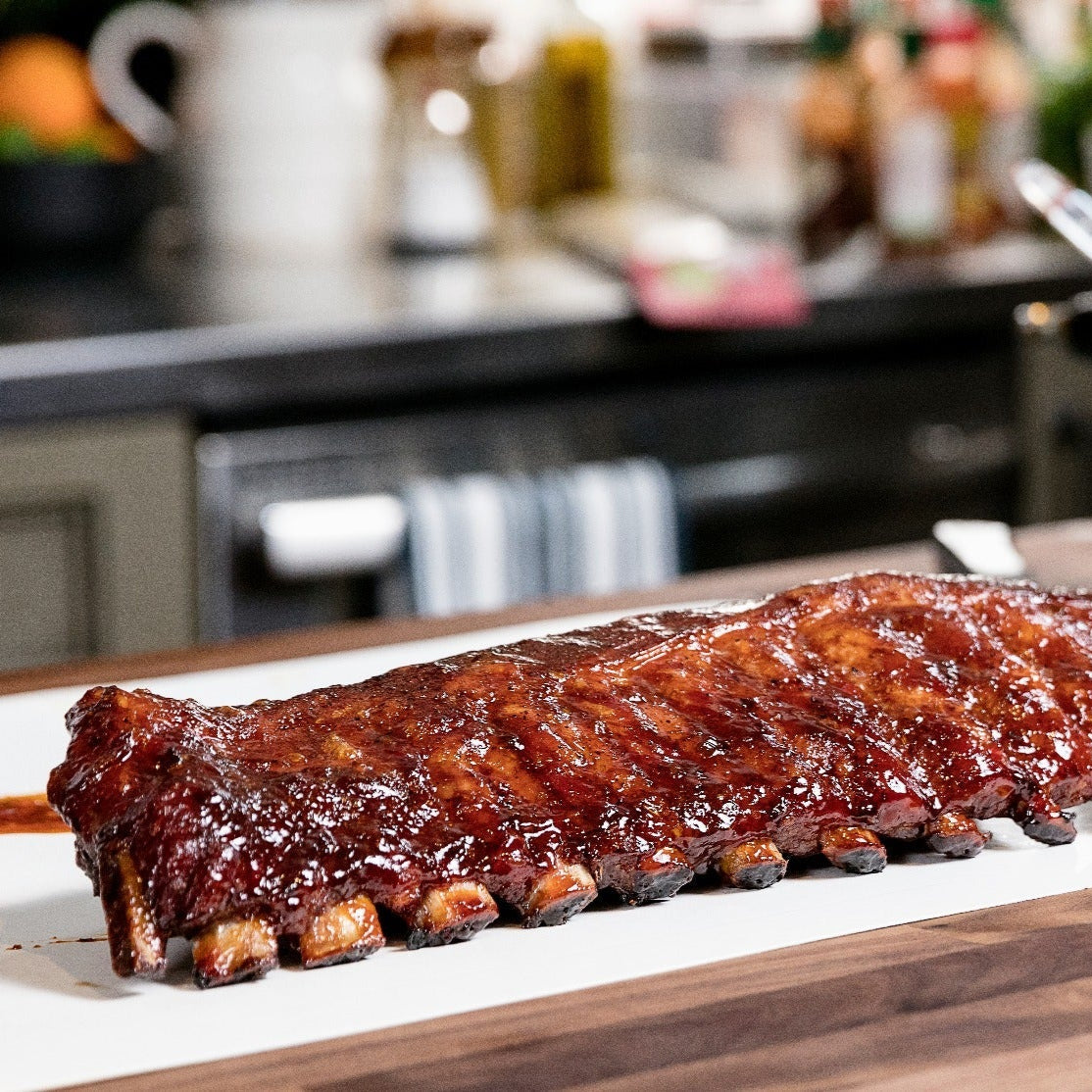 Glazed barbecue ribs placed on a white serving board in a modern kitchen setting. The ribs are richly coated in a shiny barbecue sauce, with the kitchen background subtly blurred to focus attention on the deliciously prepared meat.