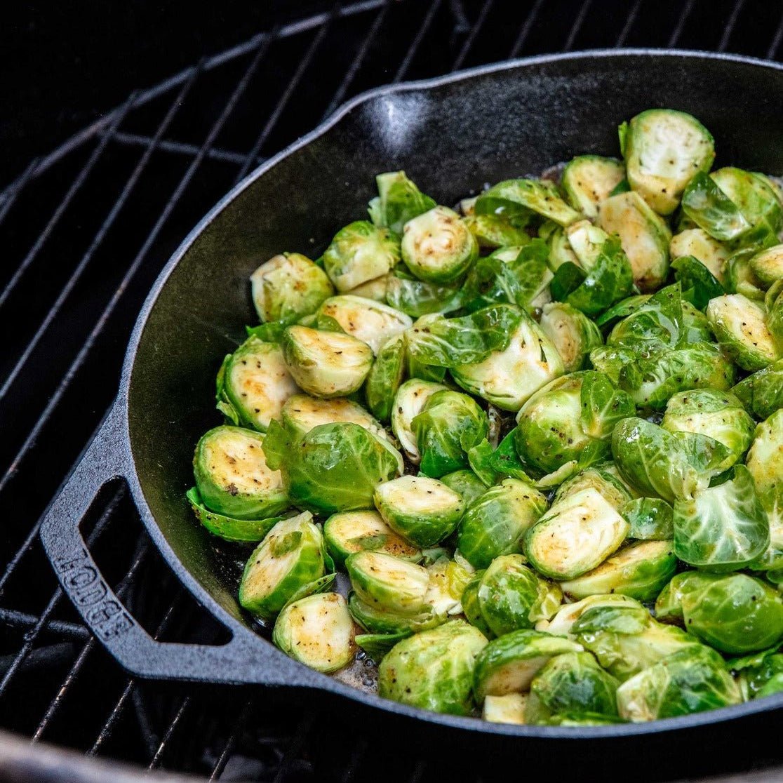 A cast iron skillet filled with halved Brussels sprouts, lightly seasoned, and cooking on a grill. The vibrant green sprouts are slightly browned on the edges, indicating they are being grilled to perfection.