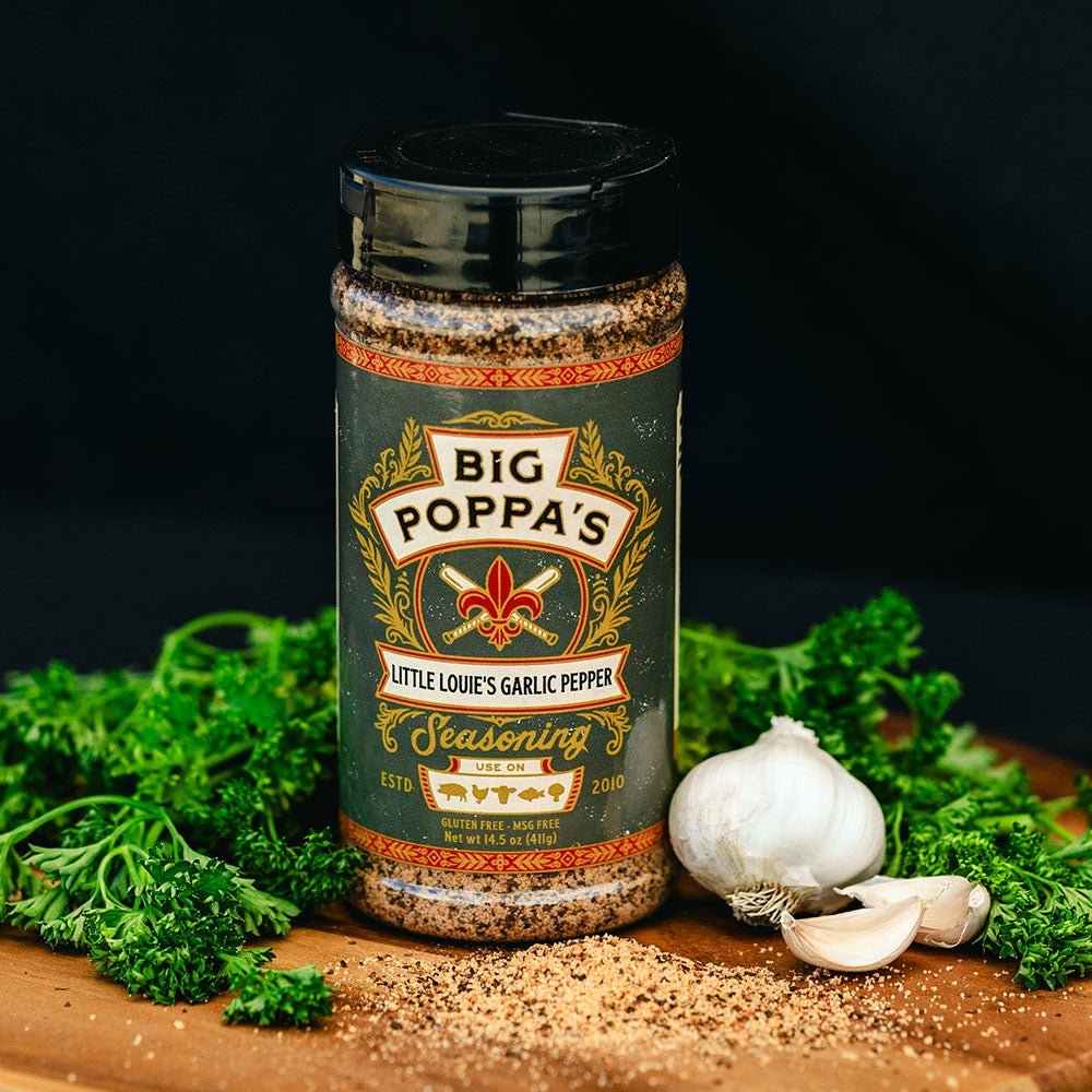 A jar of Big Poppa's Little Louie's Garlic Pepper Seasoning placed on a wooden board surrounded by fresh parsley and whole garlic bulbs. The seasoning jar has a black lid and a decorative label, and is displayed against a dark background.