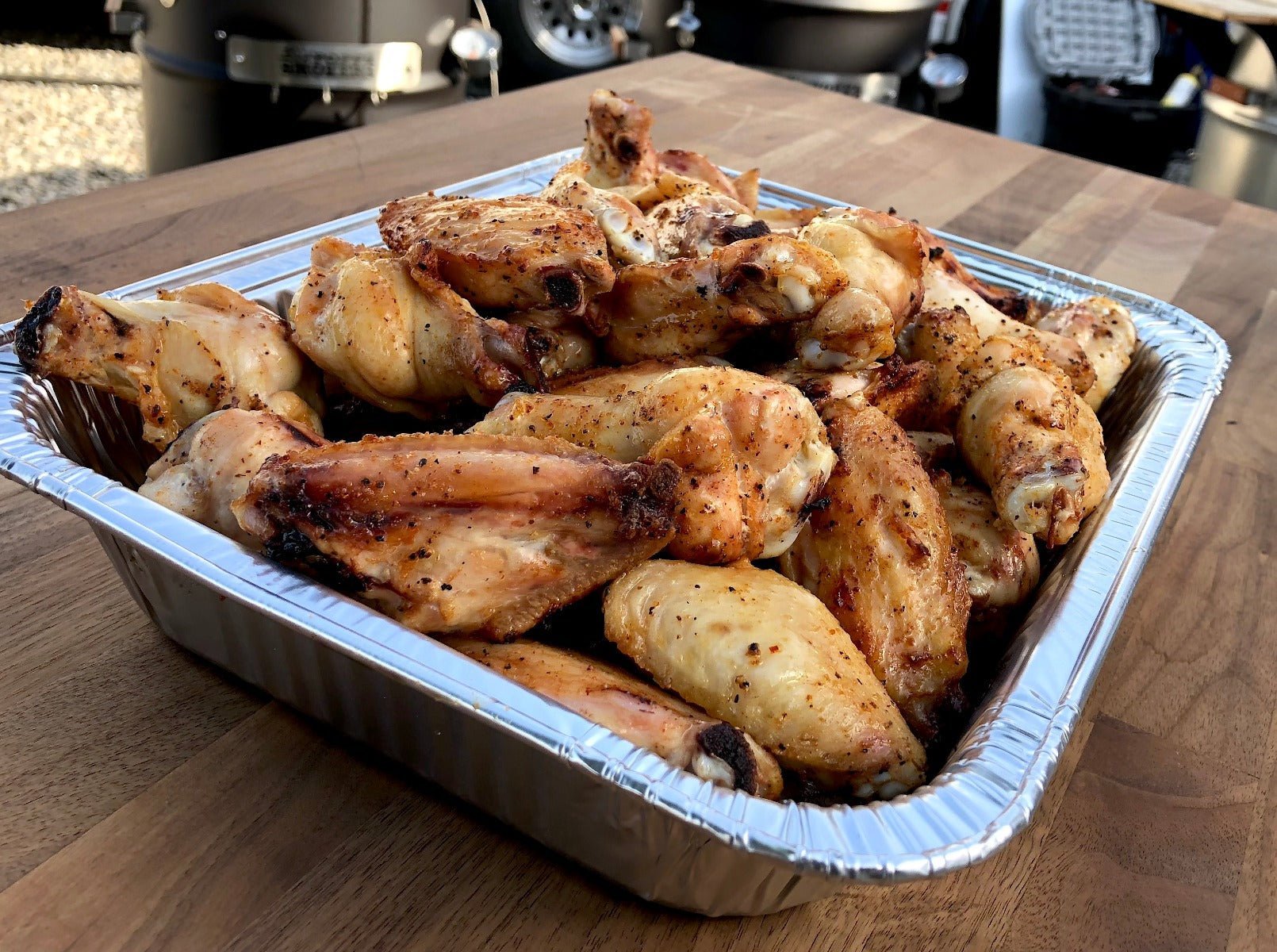 A disposable aluminum tray filled with grilled chicken wings, seasoned and cooked to a golden brown. The tray is placed on a wooden surface with outdoor grilling equipment visible in the background.
