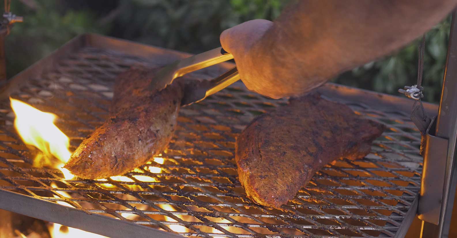 Two steaks being grilled over an open flame, with a person using tongs to turn one steak, showcasing the outdoor cooking process and the steaks' seasoned crust.