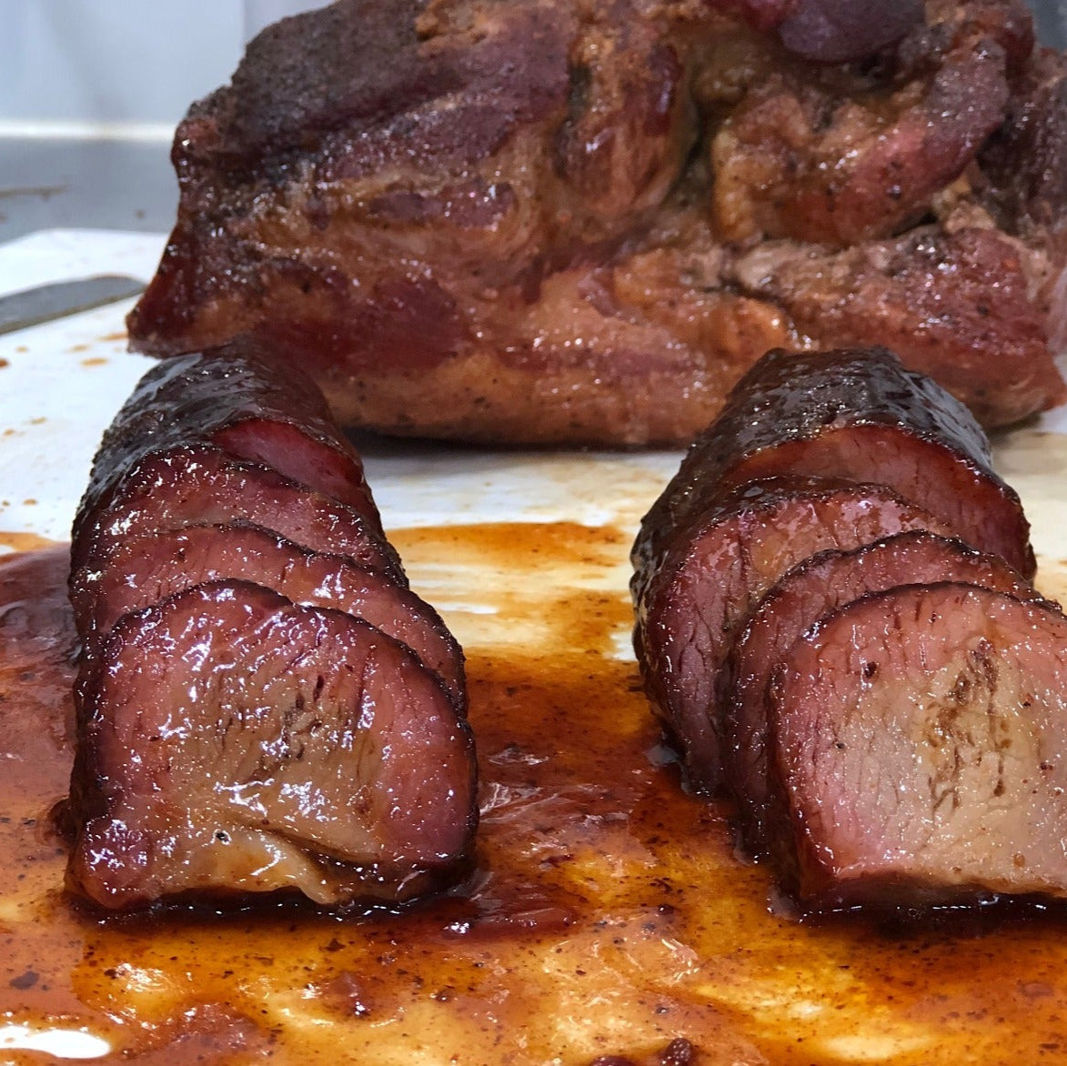 Three pieces of barbecued meat sliced and displayed on a metal tray, glazed and shiny, next to a larger chunk of similarly seasoned meat, demonstrating the application of seasoning in a savory, cooked state.