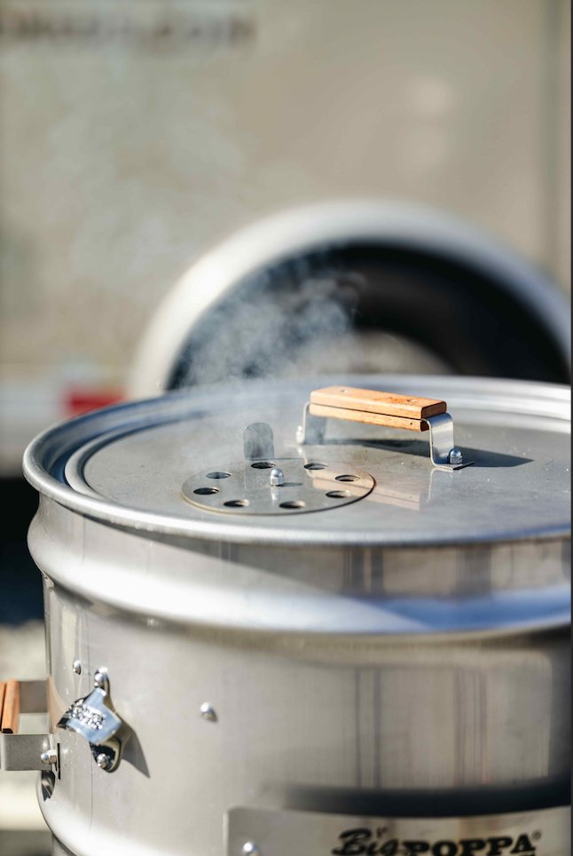 A close-up of a shiny metal smoker with a wooden handle on its lid, emitting a small amount of smoke. The brand name "Big Poppa Smokers" is visible on the side of the smoker.