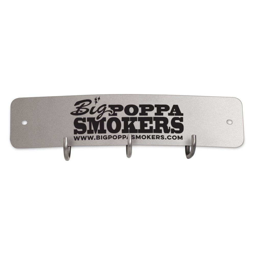 A rectangular metal tool hook shield featuring the logo of Big Poppa Smokers. The shield has a silver-grey finish with the logo in black, including the website 'www.bigpoppasmokers.com' printed below. It includes three hooks for hanging tools, centrally located along the bottom edge.