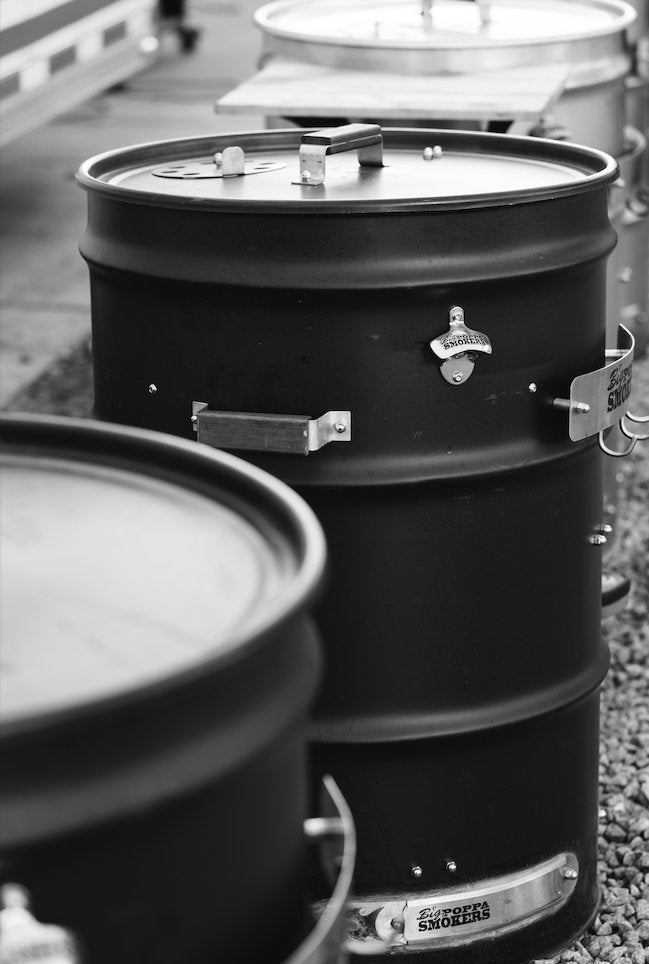 A row of black metal barrel smokers with "Big Poppa Smokers" branding are lined up on a gravel surface. The image is in black and white, focusing on the detailed craftsmanship of the smokers.