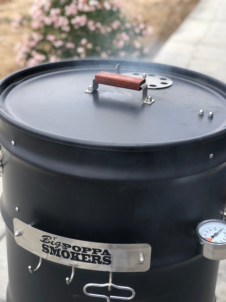 Close-up of a black barrel smoker with a wooden handle on the lid, displaying the "Big Poppa Smokers" logo. Smoke is visible rising from the smoker, with blurred flowers in the background.