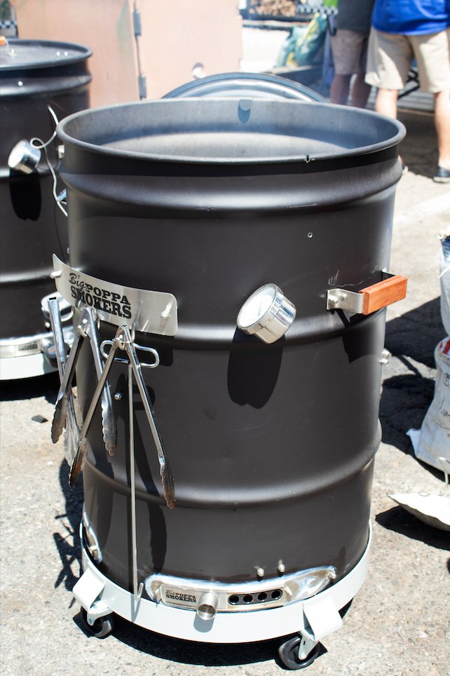 A black barrel smoker with a thermometer and grilling tools attached to the side. The smoker is in use, with smoke visible, and the "Big Poppa Smokers" logo is prominently displayed.