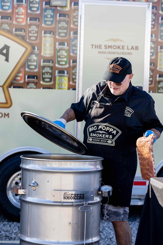 An elderly man wearing a black apron and hat with "Big Poppa Smokers" logo is opening the lid of a large metal smoker. He holds a seasoned rack of ribs in one hand, preparing to place it inside the smoker. In the background, a trailer is decorated with images of various seasoning bottles and the text "The Smoke Lab".