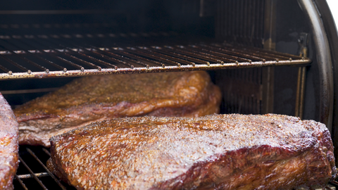 Close-up of two briskets with a crusty seasoning layer, cooking on a smoker grill with visible grill marks and a smoky background.