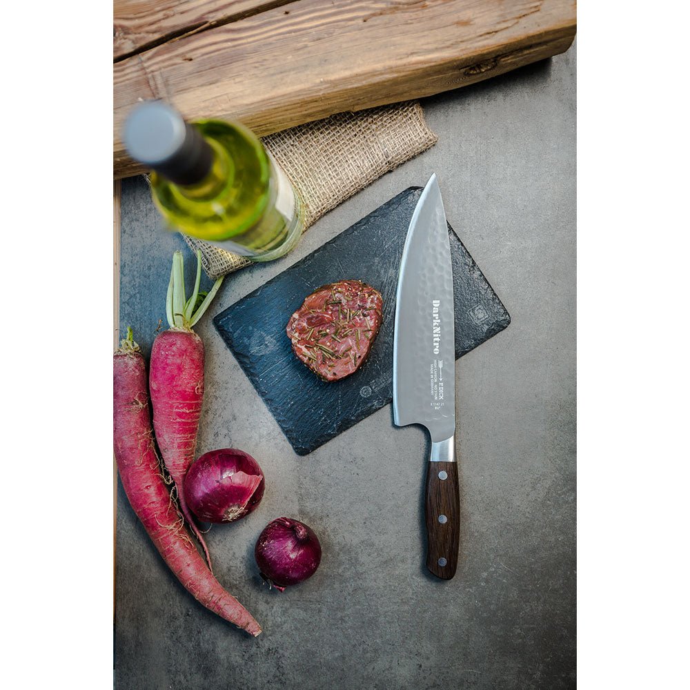 A DarkNitro chef's knife with a hammered blade finish lies on a dark stone board next to a seasoned steak. Surrounding the board are two purple carrots, two red onions, and a bottle of white wine. The setting is rustic with a wooden log in the background.