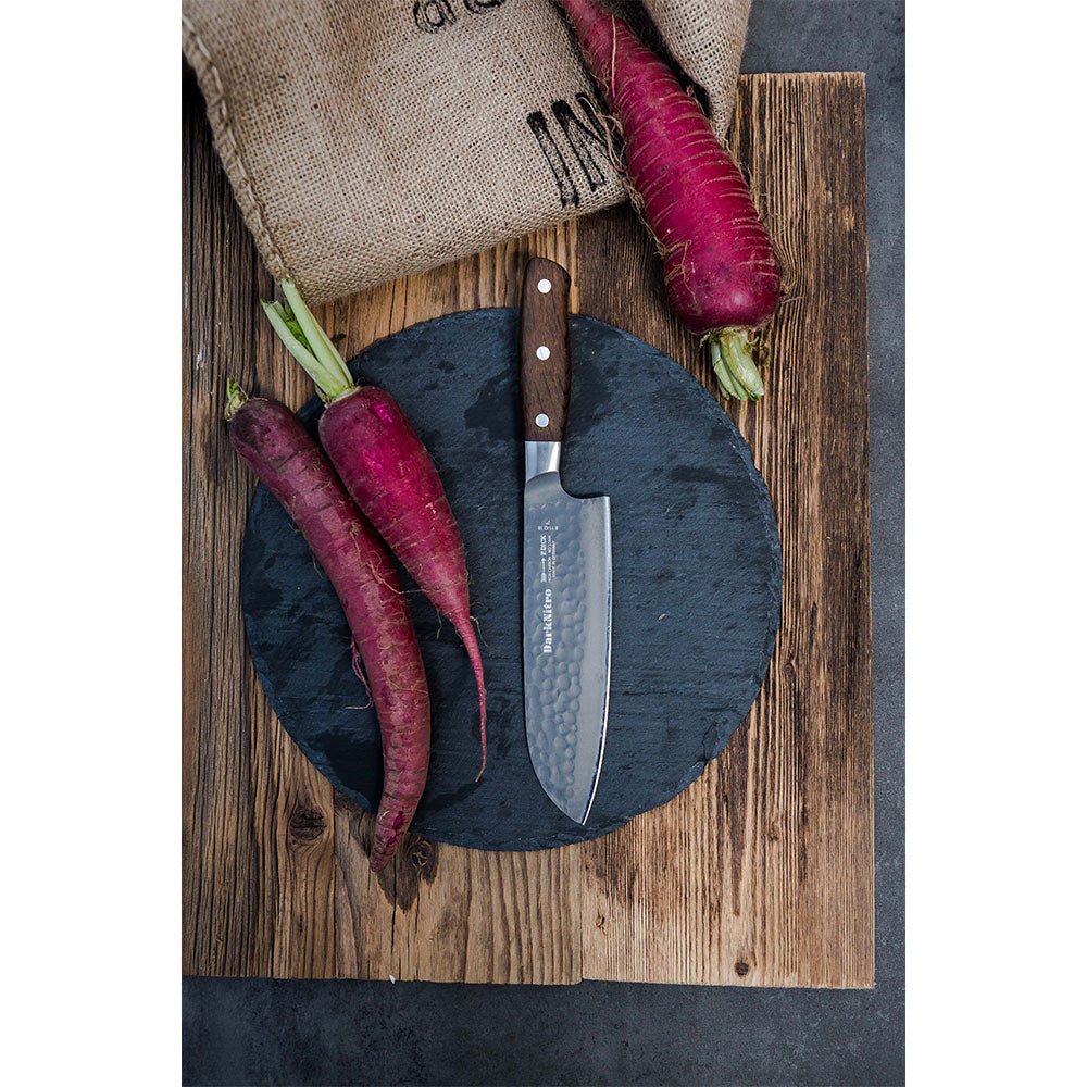 A DarkNitro Santoku knife with a hammered blade finish is displayed on a round slate serving board, surrounded by three purple carrots. A burlap sack with black text is partially visible in the background.