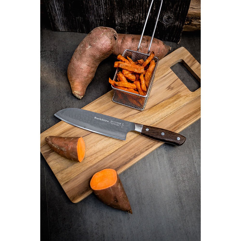 A DarkNitro Santoku knife with a hammered blade finish lies on a wooden cutting board. Surrounding the board are sweet potatoes, some whole and some cut, along with a basket of sweet potato fries.