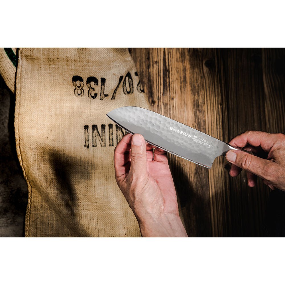 A person holds a DarkNitro Santoku knife with a hammered blade finish, showcasing the knife against a background of a burlap sack with black text. The wooden surface adds a rustic touch.