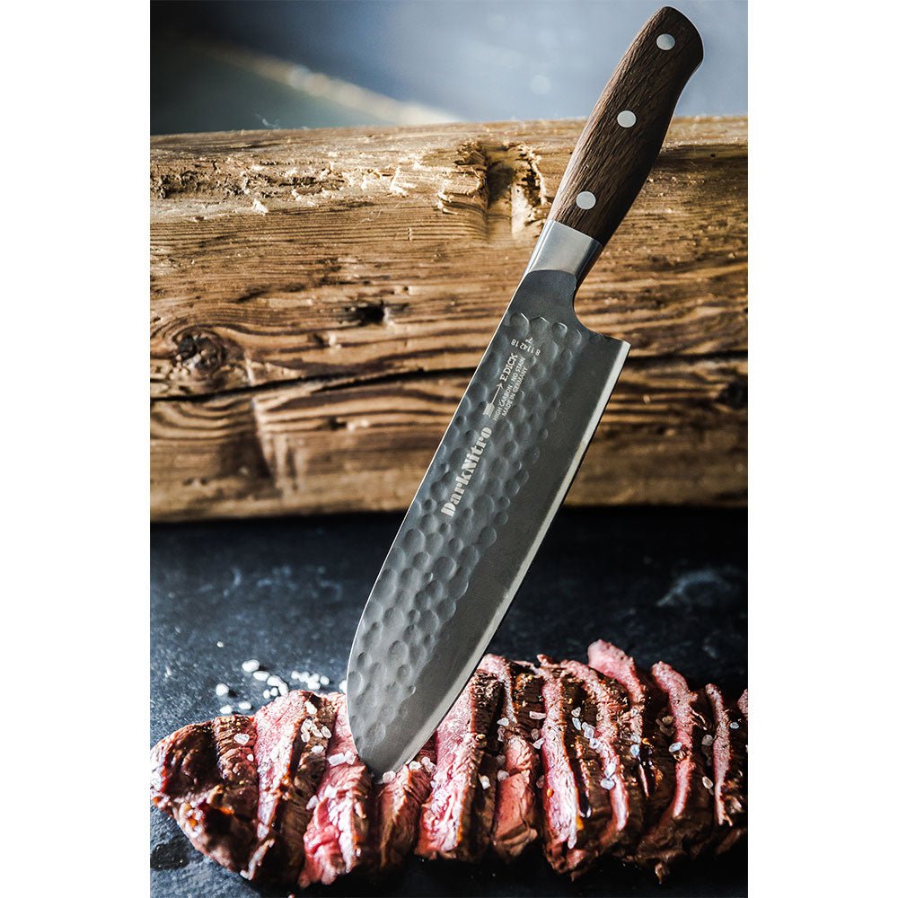 A DarkNitro Santoku knife with a hammered blade finish is shown slicing through a piece of cooked steak, placed on a dark surface with coarse salt sprinkled around.