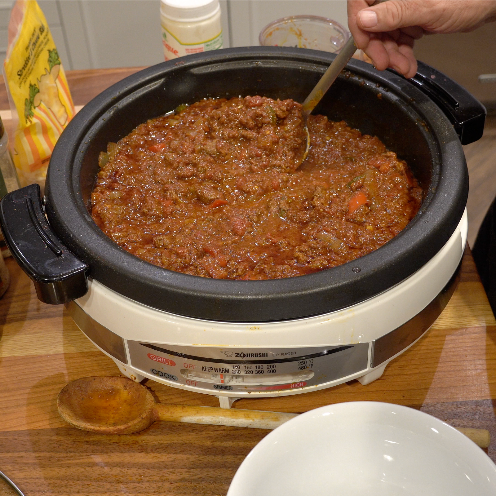 A slow cooker filled with chunky chili being stirred, surrounded by ingredients on a wooden counter.