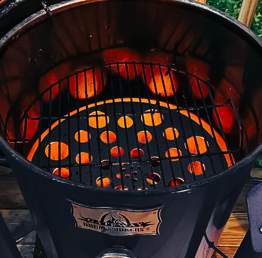 A drum smoker with a glowing fire inside, showing both the diffuser plate with holes and the cooking grate installed above it.