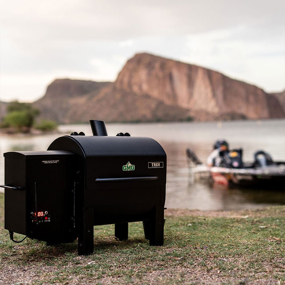 A grill placed on a grassy area near a lake with a boat in the water and mountains in the background.