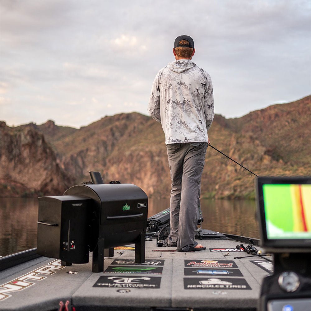 A person standing on a boat, facing away, with a mountainous landscape in the background. The boat has a grill and various stickers on the deck.