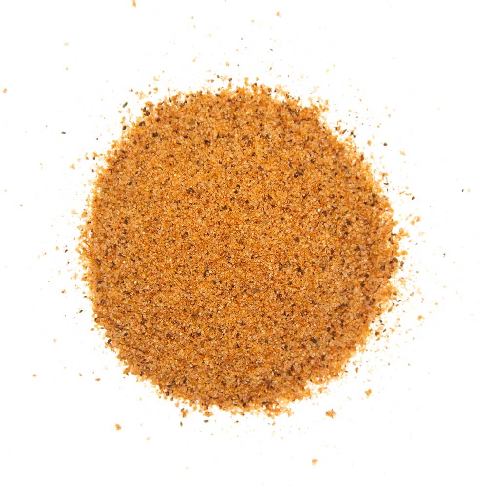 Close-up view of a pile of fine, brownish seasoning blend with visible granules of salt and spices, isolated on a white background, suggesting a texture-rich composition.