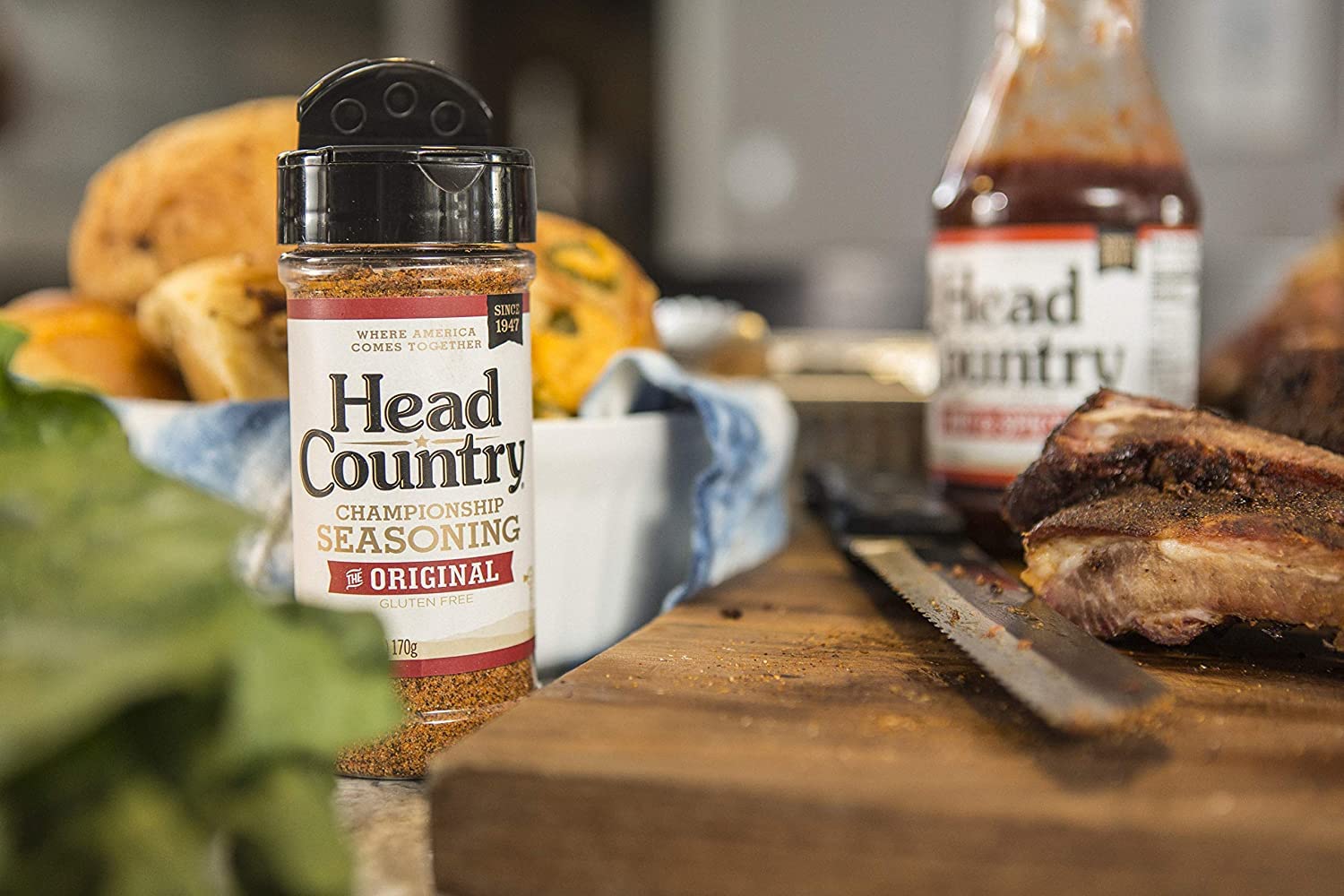 A bottle of Head Country Championship Seasoning placed prominently in the foreground on a wooden surface, surrounded by freshly baked bread, a knife, and ribs. In the background, another bottle of Head Country sauce is visible.