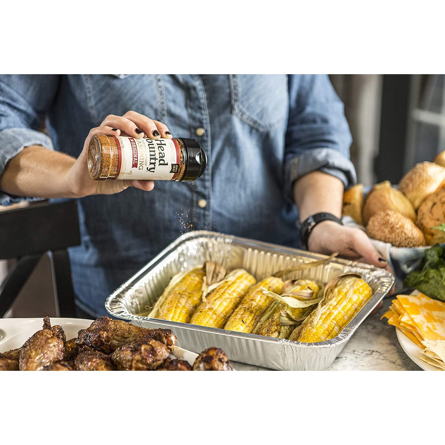 A person in a denim shirt sprinkling Head Country Championship Seasoning onto ears of corn in a foil tray. Surrounding the tray are grilled chicken pieces, bread rolls, and slices of cheese.