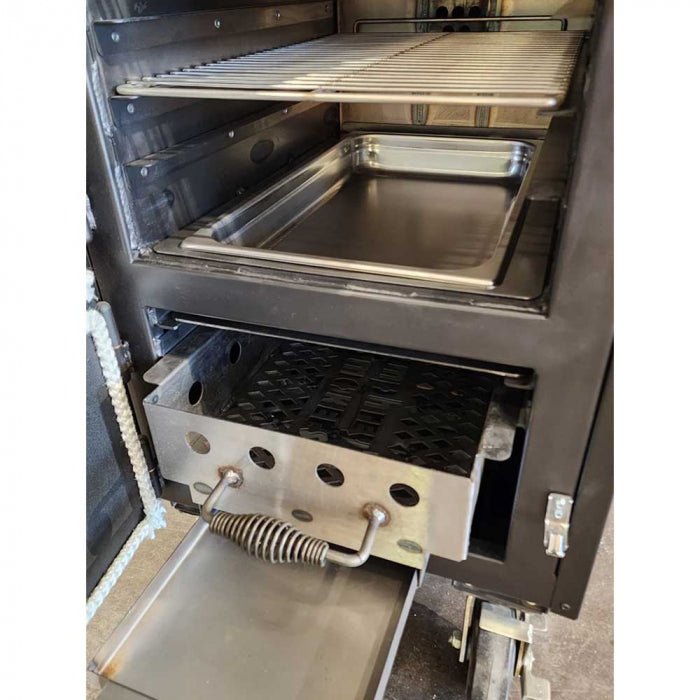 Close-up of the interior of a BBQ smoker with an open door, showing metal racks, a drip pan, and a charcoal drawer with a spring handle at the bottom.