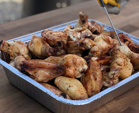 Grilled chicken wings in a foil pan, golden-brown and seasoned, with some wings showing char marks, suggesting they are freshly cooked.