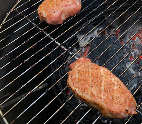 Two seasoned cuts of meat cooking on a grill, showing grill marks with glowing coals beneath, highlighting a backyard barbecuing scene.