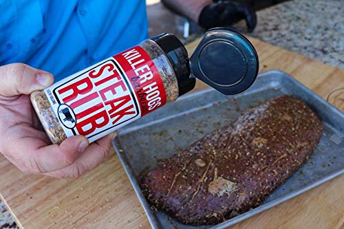 A person holds a bottle of Killer Hogs Steak Rub, sprinkling the seasoning onto a raw steak on a metal tray. The label on the bottle is red and white, reading "Killer Hogs Steak Rub."