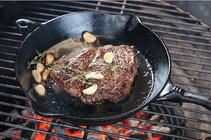 A grilled steak sits in a cast iron skillet on a barbecue grill. The steak is garnished with garlic cloves and fresh herbs, with visible grill flames underneath.