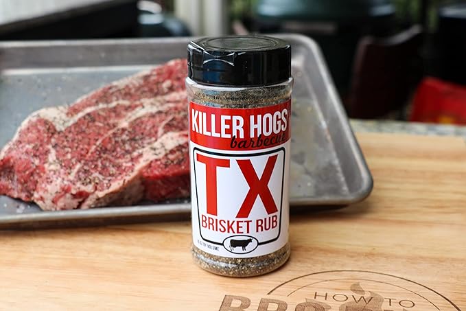 A bottle of Killer Hogs TX Brisket Rub on a wooden cutting board next to a raw, seasoned brisket on a metal tray. The bottle features a red and white label with the product name prominently displayed.