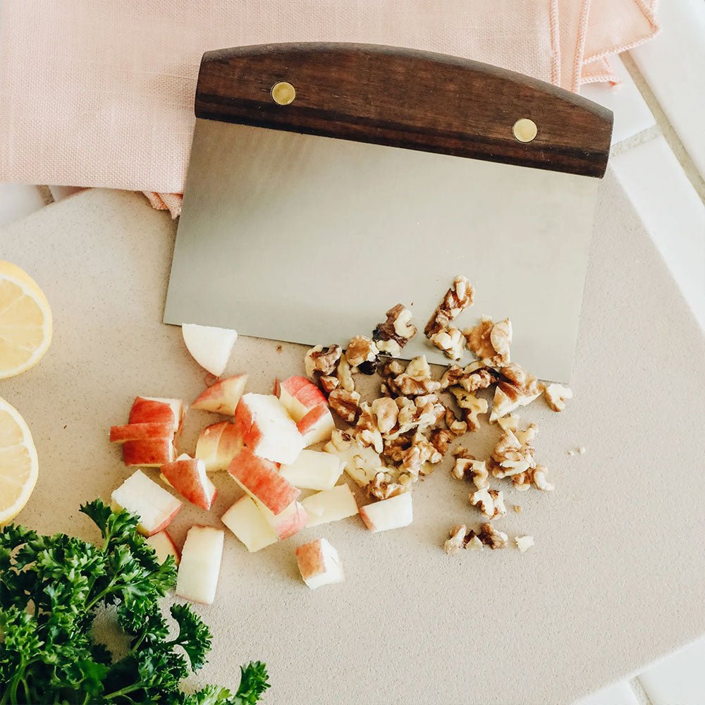 A metal dough scraper with a wooden handle is shown on a cutting board, alongside chopped apples and walnuts. Fresh parsley and lemon halves are visible on the left side, with a light pink cloth in the background.