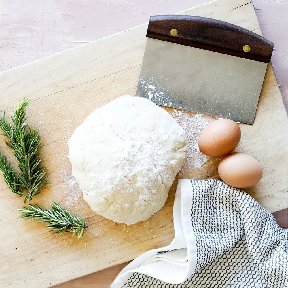 Cutting board with dough, eggs, rosemary, and dough scraper on checkered cloth.