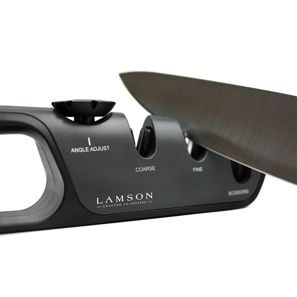 A close-up view of a Lamson adjustable angle knife sharpener with a knife being sharpened in the coarse slot. The sharpener features labeled slots for angle adjustment, coarse sharpening, fine sharpening, and scissors.