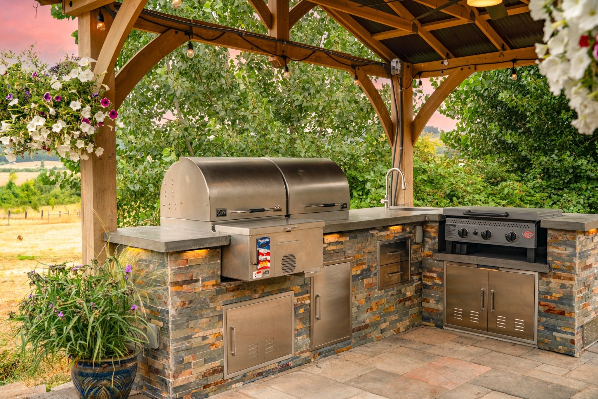 Side view of an outdoor kitchen with a MAK Grills pellet smoker, built-in grill, and a sink, framed by hanging flowers and a wooden pergola.