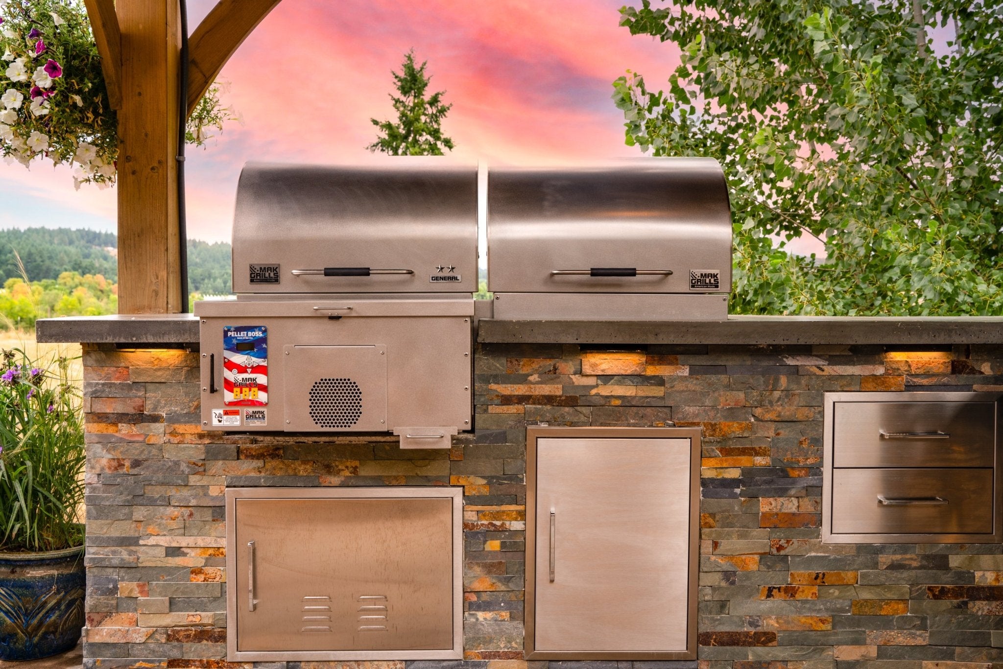 Outdoor kitchen setup with a stainless steel MAK Grills pellet smoker and additional grill equipment, set against a backdrop of trees and a vibrant sunset.