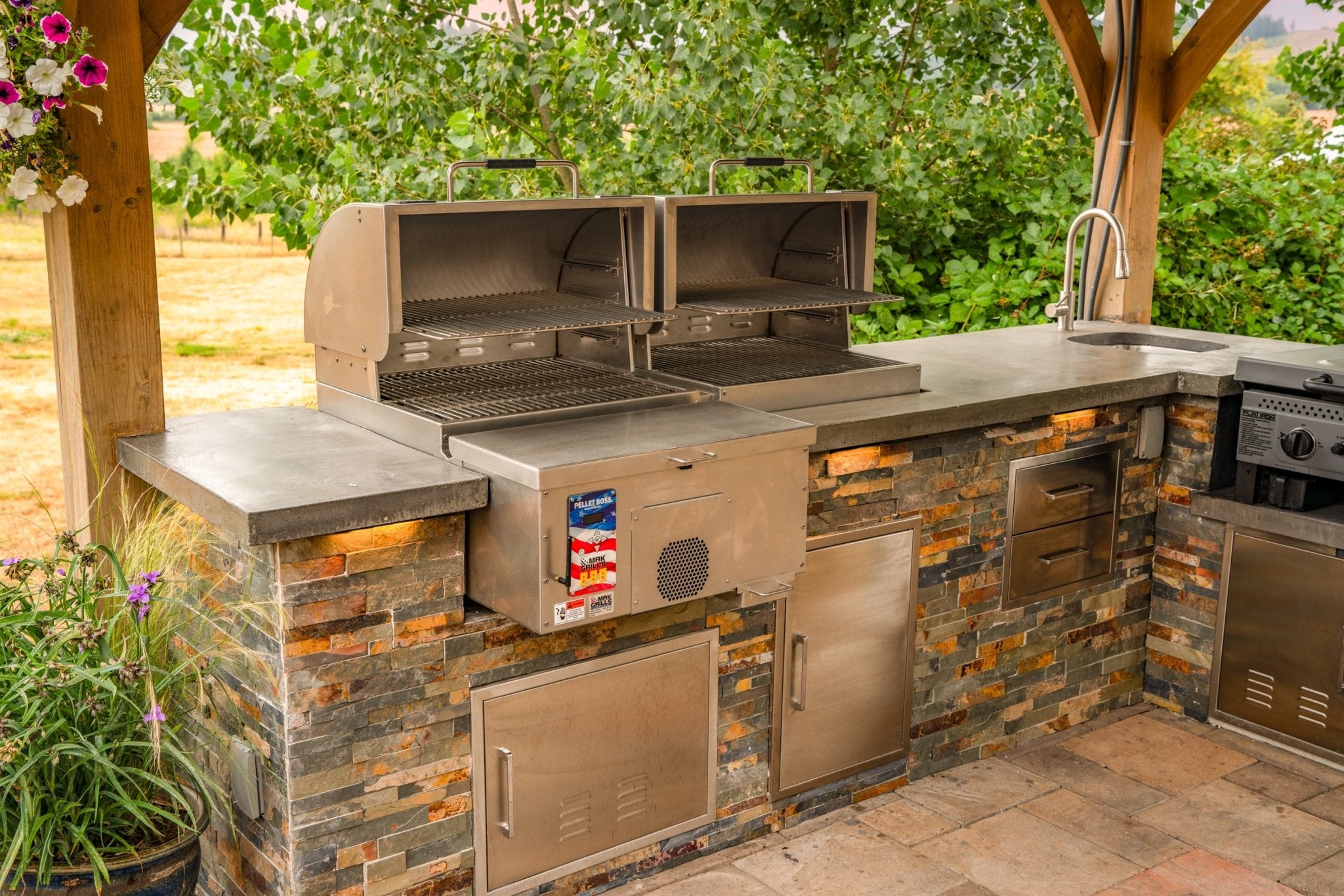 Outdoor kitchen with open MAK Grills pellet smoker and grill, under a wooden pergola, surrounded by flowers and greenery.