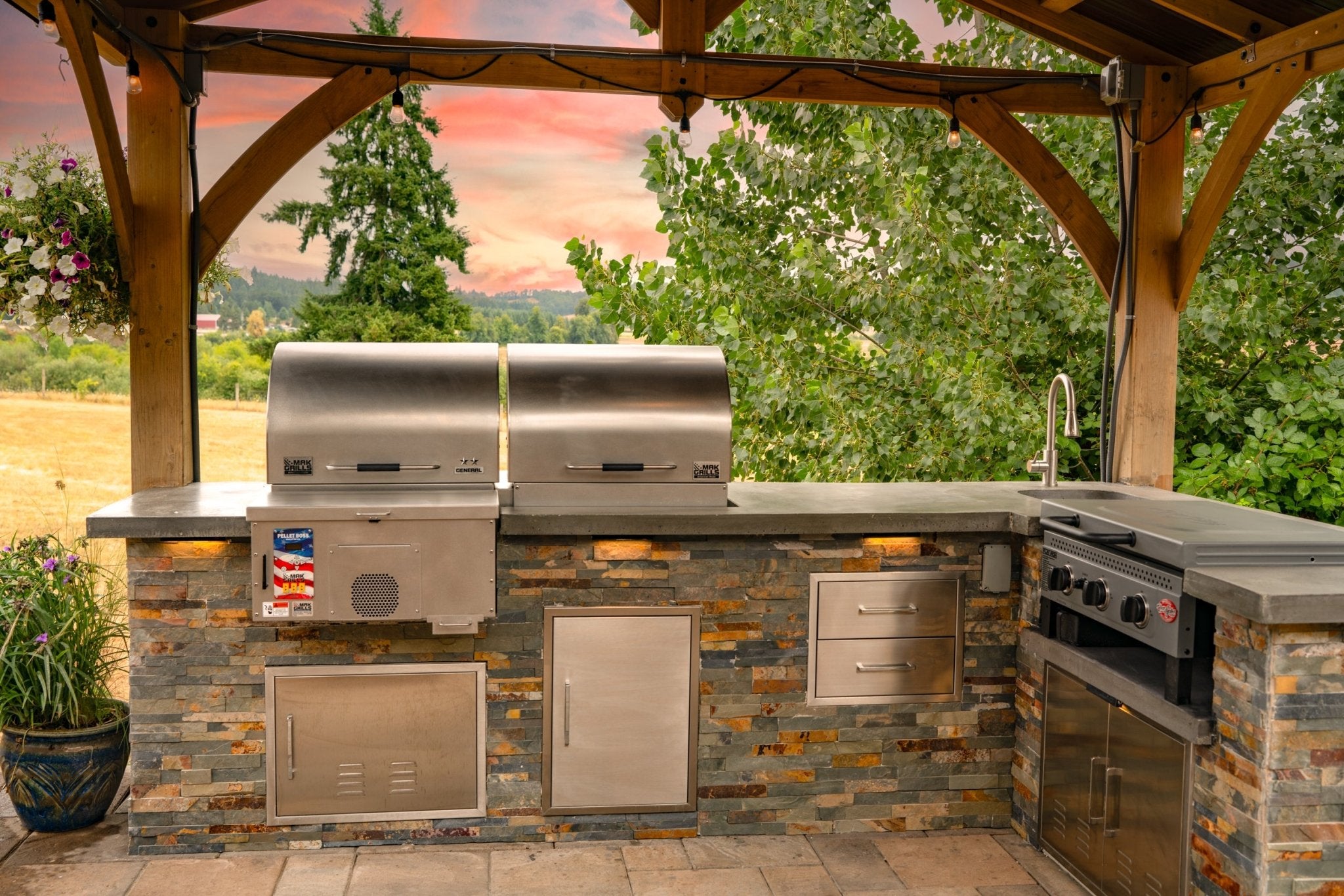 Front view of a MAK Grills pellet smoker in an outdoor kitchen, with a scenic background of trees and a vibrant sunset.