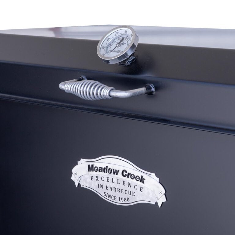 Close-up view of the Meadow Creek BBQ42 Chicken Cooker showing the thermometer and the handle on the lid. The cooker features the "Meadow Creek Excellence in Barbecue Since 1980" logo.