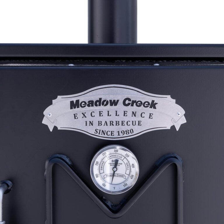 Close-up of the thermometer and logo on the upper door of the Meadow Creek BX25 smoker, displaying the text 'Meadow Creek Excellence in Barbecue Since 1980' above the temperature gauge.