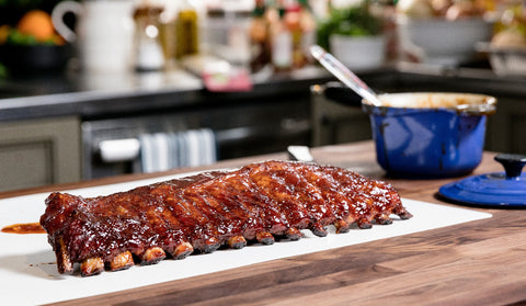 Glazed barbecue ribs placed on a white serving board in a modern kitchen setting. The ribs are richly coated in a shiny barbecue sauce, with the kitchen background subtly blurred to focus attention on the deliciously prepared meat.