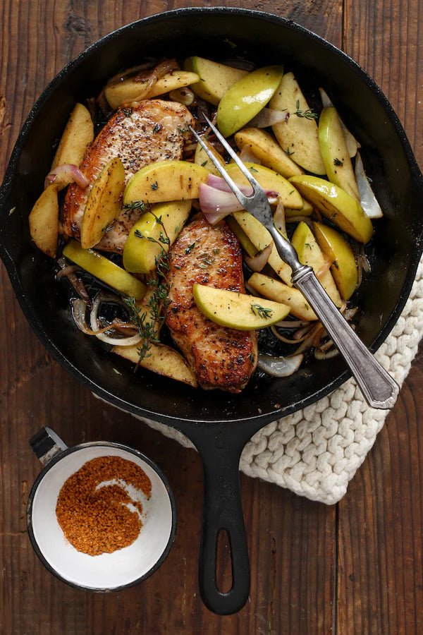 A black skillet filled with cooked pork chops, green apple slices, onions, and sprigs of thyme, with a cup of Tuffy Stone Classic BBQ Rub Sweet Heat seasoning on the side. The skillet rests on a wooden surface with a white knit cloth underneath.