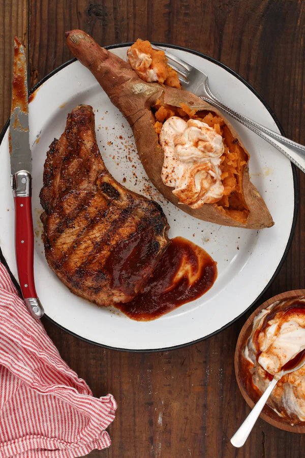 A plate with a grilled pork chop seasoned with Tuffy Stone Everything Seasoning, served with a baked sweet potato topped with a dollop of spiced sour cream. A red-handled knife is placed on the left side of the plate, which is on a wooden table with a red-striped napkin.